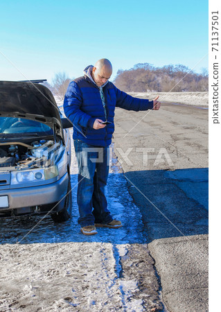 Upset young man hitchhiking and dialing for help standing near a broken car at winter roadside Upset young man hitchhiking and dialing for help standing near a broken car at winter roadside 71137501