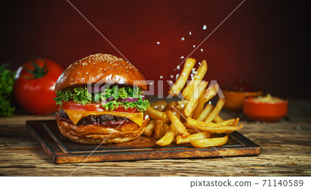 French fries fall next to cheeseburger, lying on vintage wooden cutting board. 71140589