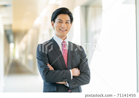 站立在他的胳膊的辦公室走廊的一個年輕微笑的人被交叉 站立在他的胳膊的辦公室走廊的一個年輕微笑的人被交叉 71141450
