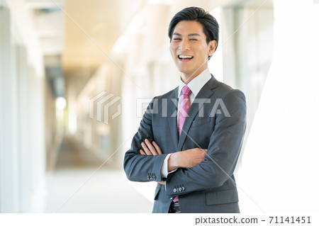 站立在他的胳膊的辦公室走廊的一個年輕微笑的人被交叉 站立在他的胳膊的辦公室走廊的一個年輕微笑的人被交叉 71141451