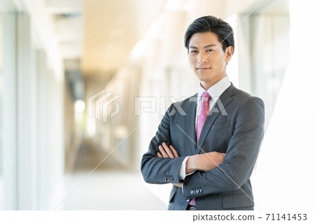 站立在他的胳膊的辦公室走廊的一個年輕微笑的人被交叉 站立在他的胳膊的辦公室走廊的一個年輕微笑的人被交叉 71141453