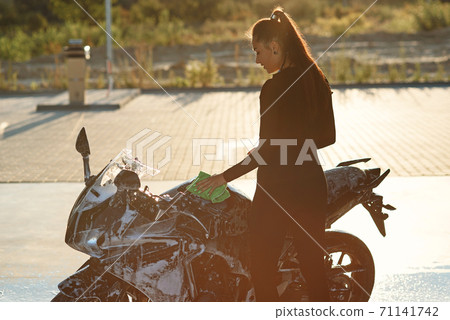Seductive young woman washing stylish sport motorcycle and wipes it from purple foam. Taking care about vehicle. Seductive young woman washing stylish sport motorcycle and wipes it from purple foam. Taking care about vehicle. 71141742