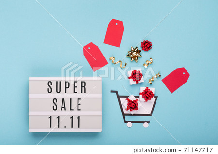 11.11 Super Sale text on white lightbox, shopping trolley, gift boxes and red tags on blue paper background. 71147717