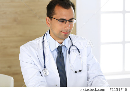 Doctor man typing at laptop computer while sitting at the desk at his working place. Perfect medical service in clinic. Data in medicine and healthcare Doctor man typing at laptop computer while sitting at the desk at his working place. Perfect medical service in clinic. Data in medicine and healthcare 71151746