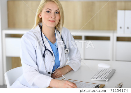 Doctor woman at work while sitting at the desk in hospital or clinic. Blonde cheerful physician filling up medical records form. Data and best service in medicine and healthcare 71154174