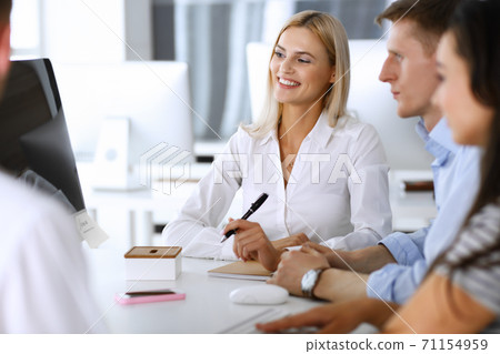 Group of business people discussing questions at meeting. Headshot of blonde businesswoman while smiling to her colleague at office negotiation. Teamwork and cooperation in corporate occupation 71154959