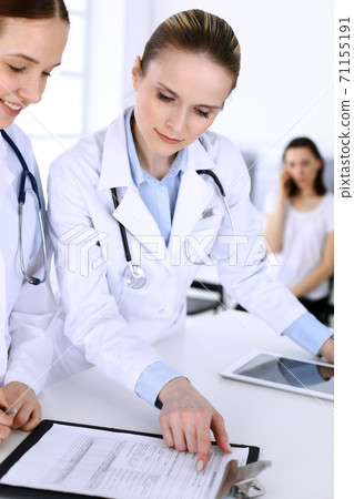 Group of doctors at work in hospital with patient in a queue at the background. Physician filling up medical documents or prescription while standing at reception desk. Data in medicine and health Group of doctors at work in hospital with patient in a queue at the background. Physician filling up medical documents or prescription while standing at reception desk. Data in medicine and health 71155191