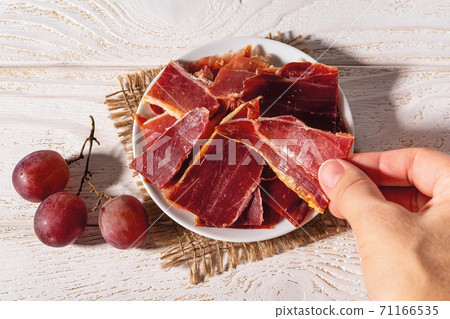 Woman hand takes a slice of jamon from a plate over white wood table. Woman hand takes a slice of jamon from a plate over white wood table. 71166535