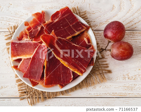 Sliced jamon on a plate and two large pink grapes over white wood table. 71166537