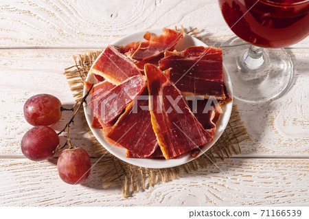Sliced jamon on a plate, few pink grapes and wine glass over white wood table. 71166539