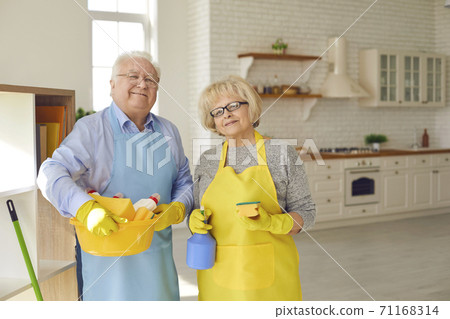 Elderly happy couple in aprons and gloves standing and holding equipment for cleaning home 71168314
