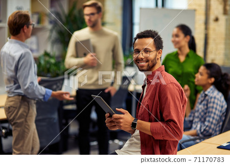 Young cheerful mixed race man holding digital tablet and smiling at camera while working with colleagues in the modern office, selective focus Young cheerful mixed race man holding digital tablet and smiling at camera while working with colleagues in the modern office, selective focus 71170783