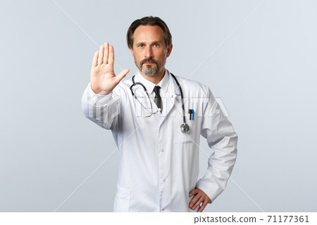 Covid-19, coronavirus outbreak, healthcare workers and pandemic concept. Serious professional doctor in white coat extend arm to show stop, making warning or prohibition gesture Covid-19, coronavirus outbreak, healthcare workers and pandemic concept. Serious professional doctor in white coat extend arm to show stop, making warning or prohibition gesture 71177361