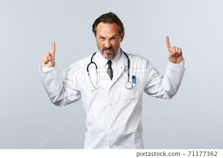Covid-19, coronavirus outbreak, healthcare workers and pandemic concept. Angry outraged male doctor in white coat, pointing fingers up grimacing furious, looking mad at camera, white background Covid-19, coronavirus outbreak, healthcare workers and pandemic concept. Angry outraged male doctor in white coat, pointing fingers up grimacing furious, looking mad at camera, white background 71177362