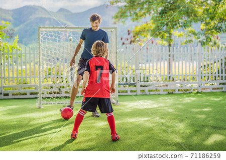 Little cute kid boy in red football uniform and his trainer or father playing soccer, football on field, outdoors. Active child making sports with kids or father, Smiling happy boy having fun in 71186259