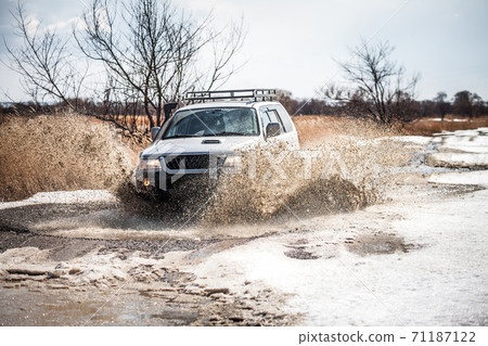 Silver SUV on dirt road in early spring making splashes from a puddle Silver SUV on dirt road in early spring making splashes from a puddle 71187122