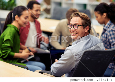Happy cheerful mature businessman smiling at camera while having brainstorming meeting with colleagues in the modern office Happy cheerful mature businessman smiling at camera while having brainstorming meeting with colleagues in the modern office 71187125