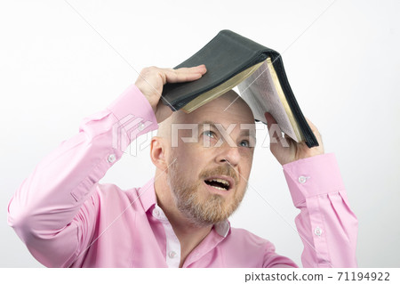 Bearded man in a pink shirt hides his head under an open Bible 71194922