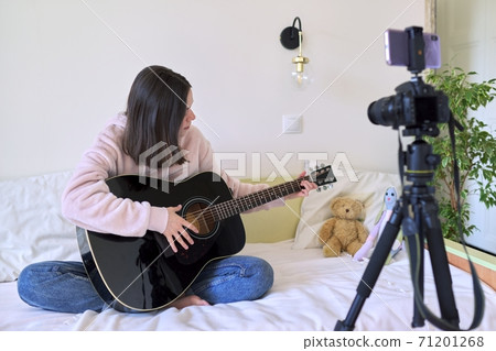 Teen girl sitting at home in bed with an acoustic guitar, girl learning to play the guitar online 71201268