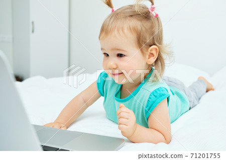 The child carefully looks into the laptop monitor while lying on the bed 71201755