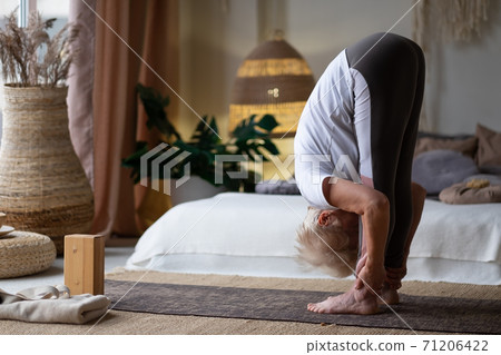 Attractive senior woman working out standing in Uttanasana at home. Attractive senior woman working out standing in Uttanasana at home. 71206422