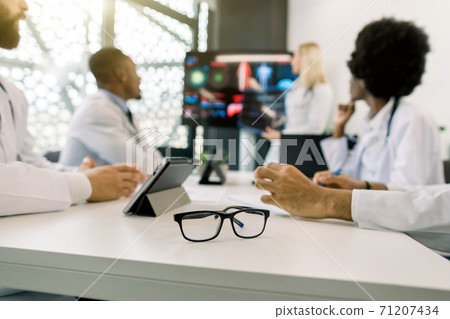 Blurred image of multiethnical concentrated medical team, having a meeting, and listening the lecture of their blond female colleague. Focus on stylish eyeglasses on the table. Education concept Blurred image of multiethnical concentrated medical team, having a meeting, and listening the lecture of their blond female colleague. Focus on stylish eyeglasses on the table. Education concept 71207434