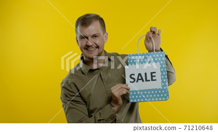 Man showing Sale word inscription with shopping bag, smiling, rejoicing discounts on Black Friday 71210648