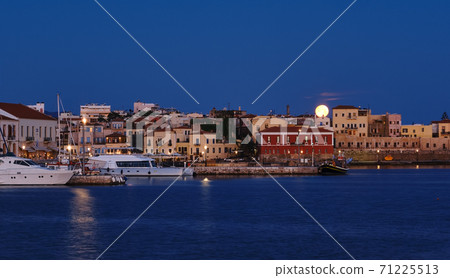 Full moon setting over Chania's Old Venetian Harbour. Night view of piers, boats and Maritime museum of Crete and houses over marina. Full moon setting over Chania's Old Venetian Harbour. Night view of piers, boats and Maritime museum of Crete and houses over marina. 71225513