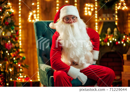 Santa sitting at the Christmas tree, near fireplace and looking at camera. Indoors Santa sitting at the Christmas tree, near fireplace and looking at camera. Indoors 71229044