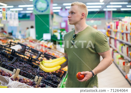 Young sporty man is choosing fruits in grocery store. Concept of healthy nutrition, balanced diet, vitamins Young sporty man is choosing fruits in grocery store. Concept of healthy nutrition, balanced diet, vitamins 71264064