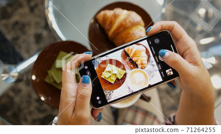 Close up of woman holds modern smartphone and taking pictures of her morning breakfast coffee, green chocolate and tasty croissant buns to share photos on social media resources 71264255
