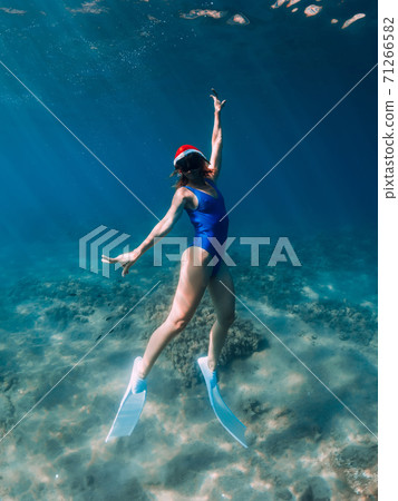 Christmas holidays party with freediver. Happy freediver woman with New year cap glides underwater with fins in blue ocean. 71266582