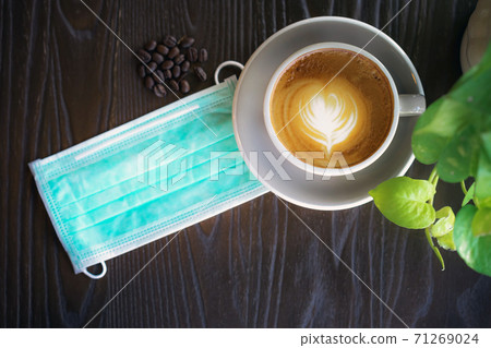 Hot Cappuccino coffee with protective mask on a wooden table for people being quarantined in the situation of the spread of the corona virus (Covid-19).  New normal lifestyle concept. Selective focus 71269024