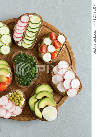 Vegetable sandwiches on the wood board on the gray background. Fresh bread with pesto, cucumber, avocado, cherry tomatoes, radish and cheese. Delicious breakfast. Flat lay, top view. 71272107