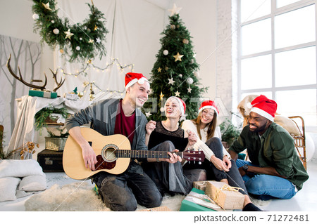 Group of friends having fun on Christmas Party. Young boy playing guitar and friends are smiling and singing. Decorated cozy room with Christmas tree. 71272481