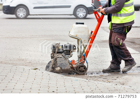 A worker cuts the old asphalt with a gas saw on the carriageway against the backdrop of a city street. 71279753