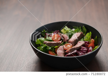 Steak salad with cherry tomatoes and mixed greens in black bowl on walnut wood background 71280872