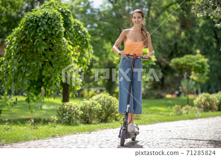 Long-haired young girl riding a scooter in the park and looking contented 71285824