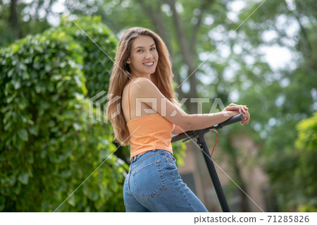 Long-haired young girl riding a scooter in the park and smiling 71285826