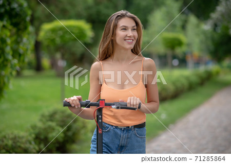 Long-haired cute girl riding scooter in the park and smiling 71285864