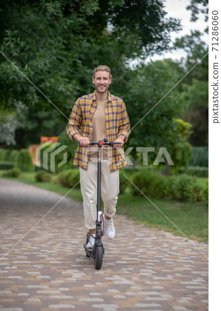 Handsome young man riding a scooter in the park and feeling good Handsome young man riding a scooter in the park and feeling good 71286060