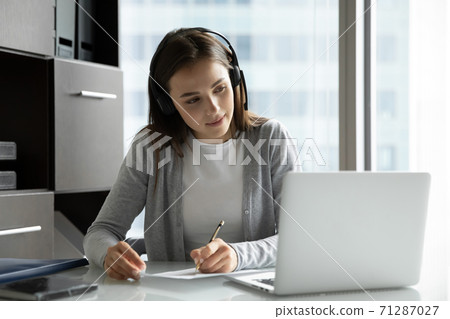 Interested woman employee watching webinar in headphones writing up information Interested woman employee watching webinar in headphones writing up information 71287027