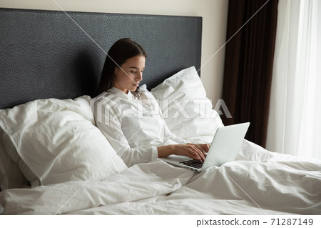Concentrated millennial woman working online at laptop in bed Concentrated millennial woman working online at laptop in bed 71287149