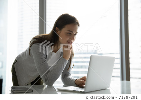 Interested female worker standing near desk looking on laptop screen 71287291
