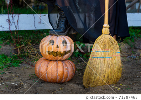 On a pumpkin with a drawn scary face is a witch's foot, next to a broom, close-up On a pumpkin with a drawn scary face is a witch's foot, next to a broom, close-up 71287465