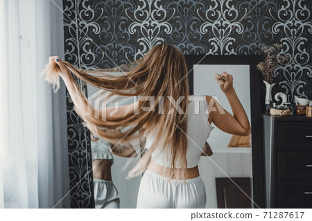 How take care of long hair. Back view of young natural blonde woman in white home clothes combing her hair and looking into the mirror in bedroom 71287617