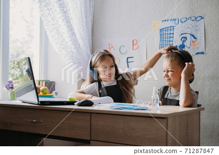 Distance learning, home education, remote lessons concept. Two schoolchildren, brother and sister in headphones sit near open laptop and watch a video lesson, listen to the teacher Distance learning, home education, remote lessons concept. Two schoolchildren, brother and sister in headphones sit near open laptop and watch a video lesson, listen to the teacher 71287847