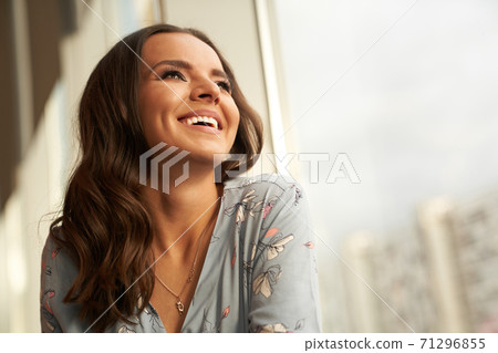 A beautiful young woman happy and smiling looks towards the window, the girl has perfect skin and a snow-white smile A beautiful young woman happy and smiling looks towards the window, the girl has perfect skin and a snow-white smile 71296855