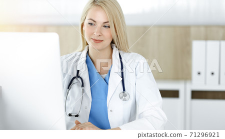 Woman-doctor at work while sitting at the desk in sunny clinic. Blonde cheerful physician using desktop computer 71296921