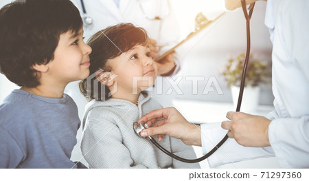 Doctor-woman examining a child patient by stethoscope in sunny clinik. Cute arab boy and his brother at physician appointment 71297360